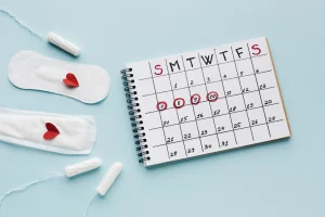 A weekly calendar with dates marked resting on a blue surface with sanitary napkins and tampons on the left of it