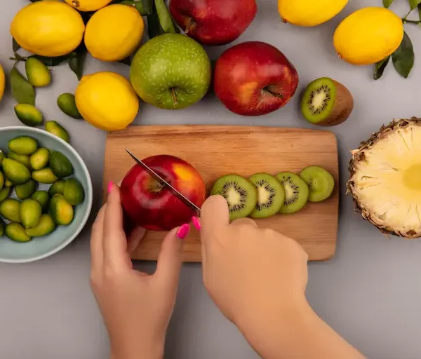 top-view-female-hands-cutting-apple-wooden-kitchen-board-with-knife-with-kinkans-bowl-with-apples-kiwi-pineapple-lemons-isolated-white-wall_Activ Living Community