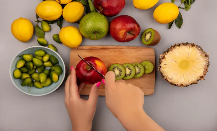 top-view-female-hands-cutting-apple-wooden-kitchen-board-with-knife-with-kinkans-bowl-with-apples-kiwi-pineapple-lemons-isolated-white-wall_Activ Living Community